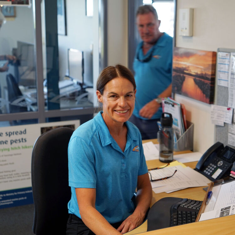 Smiling staff member at the reception desk of Tauranga Bridge Marina, with office environment and another team member in the background.
